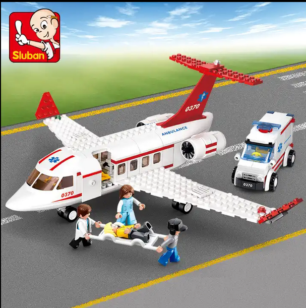 A toy medical emergency scene with a white airplane marked "Ambulance," several figures, and a small ambulance vehicle. Two figures carry a stretcher with a patient figure.